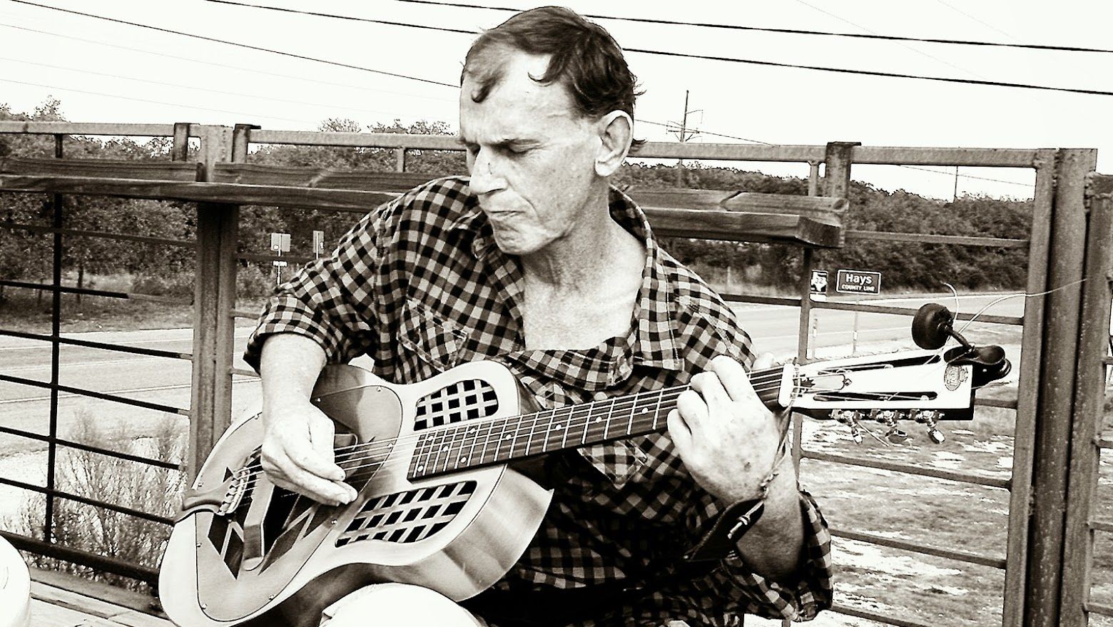 Black and white photo of a man playing a resonator guitar beside a highway overpass