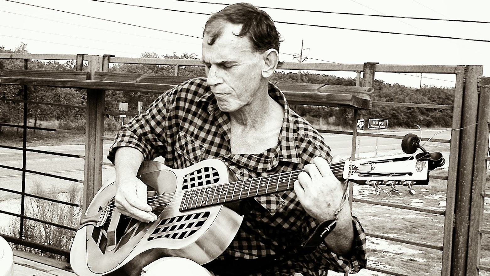 Black and white photo of a man playing a resonator guitar beside a highway overpass
