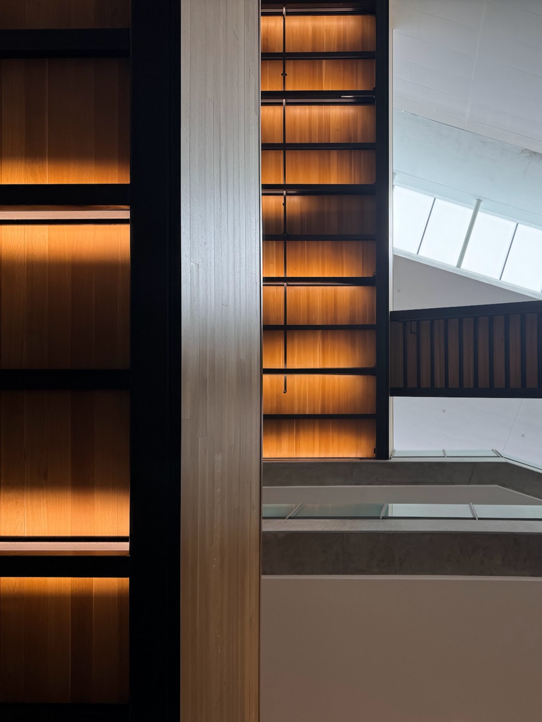 Abstract interior view of warm amber-backlit wood bookshelves beside a multi-story atrium